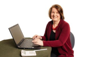 Angela Proud - Proud Bookkeeping Ltd - Director sitting at a desk with a laptop and calculator with a greeting smile